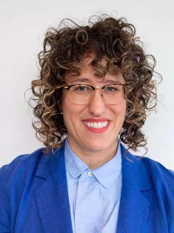 white background, woman wearing glasses, curly hair, blue blazer and light blue collared shirt