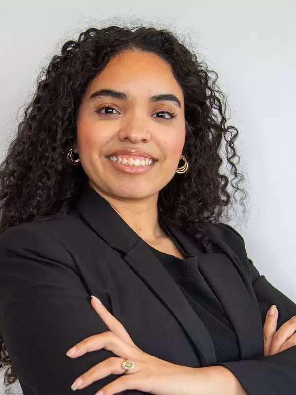 white background, woman with dark brown/black hair, wearing black blazer, smiling, arms crossed